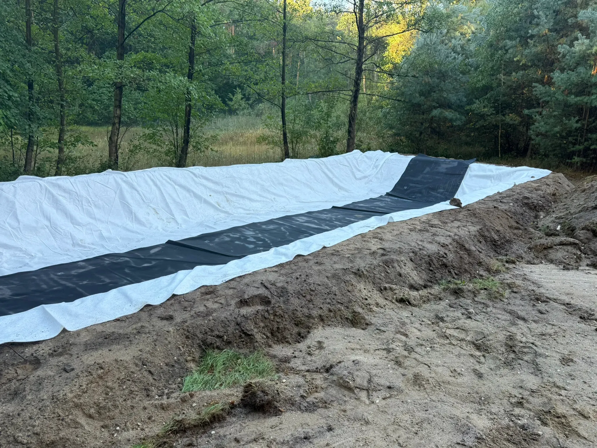 Baugrube ausgehoben und mit weißem Schutzvlies sowie schwarzer EPDM-Teichfolie ausgelegt — der Filterkörper wird gegen den umgebenden Sandboden vollständig abgedichtet, damit das durchsickernde Abwasser nur über den vorgesehenen Auslauf entweicht