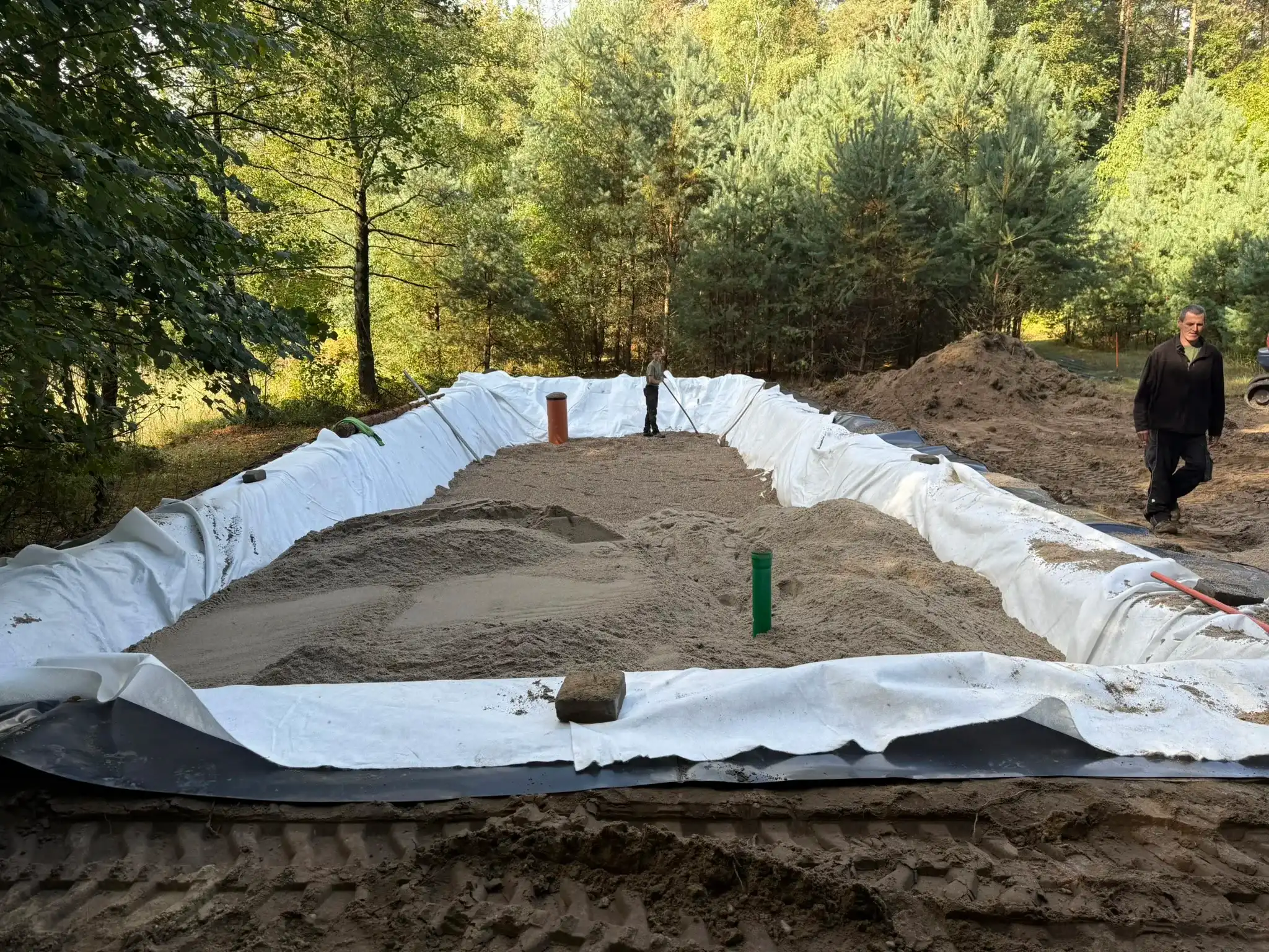Bau des Pflanzenklärbeets am Forsthaus Parnitz im September 2026 — Baugrube mit Vlies und Teichfolie ausgelegt, wird mit Filtersand gefüllt; im Hintergrund das Bauteam von Dirk Fiedler aus Bad Belzig